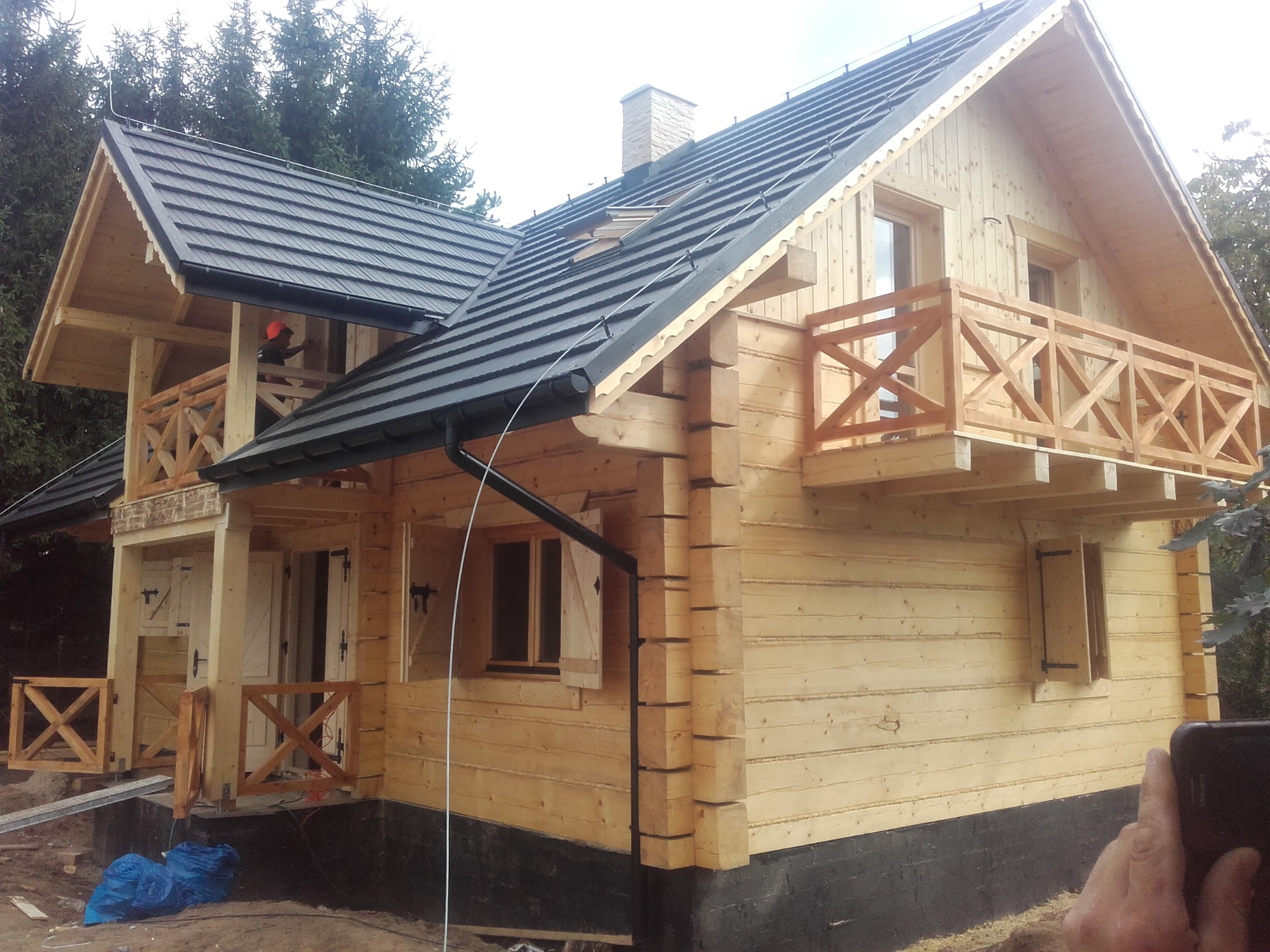 Surowy, dwukondygnacyjny dom z jasnego drewna z czarnym dachem, widoczne rusztowania i fragment osoby w pomarańczowym ubraniu na balkonie.