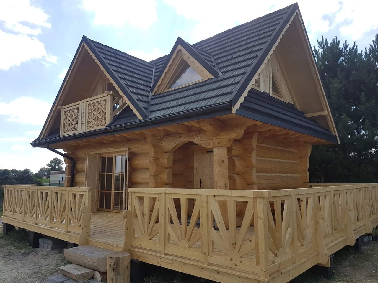 Wykończony, parterowy dom z bali w Mszanie Dolnej z drewnianą balustradą wokół tarasu i ozdobnym balkonem na poddaszu, kryty ciemną dachówką. Widoczne detale architektoniczne i naturalna faktura...