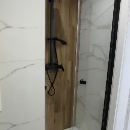 Nowoczesna kabina prysznicowa typu walk-in z czarną armaturą i panelami imitującymi drewno, wykończona białymi płytkami z marmurkowym wzorem.