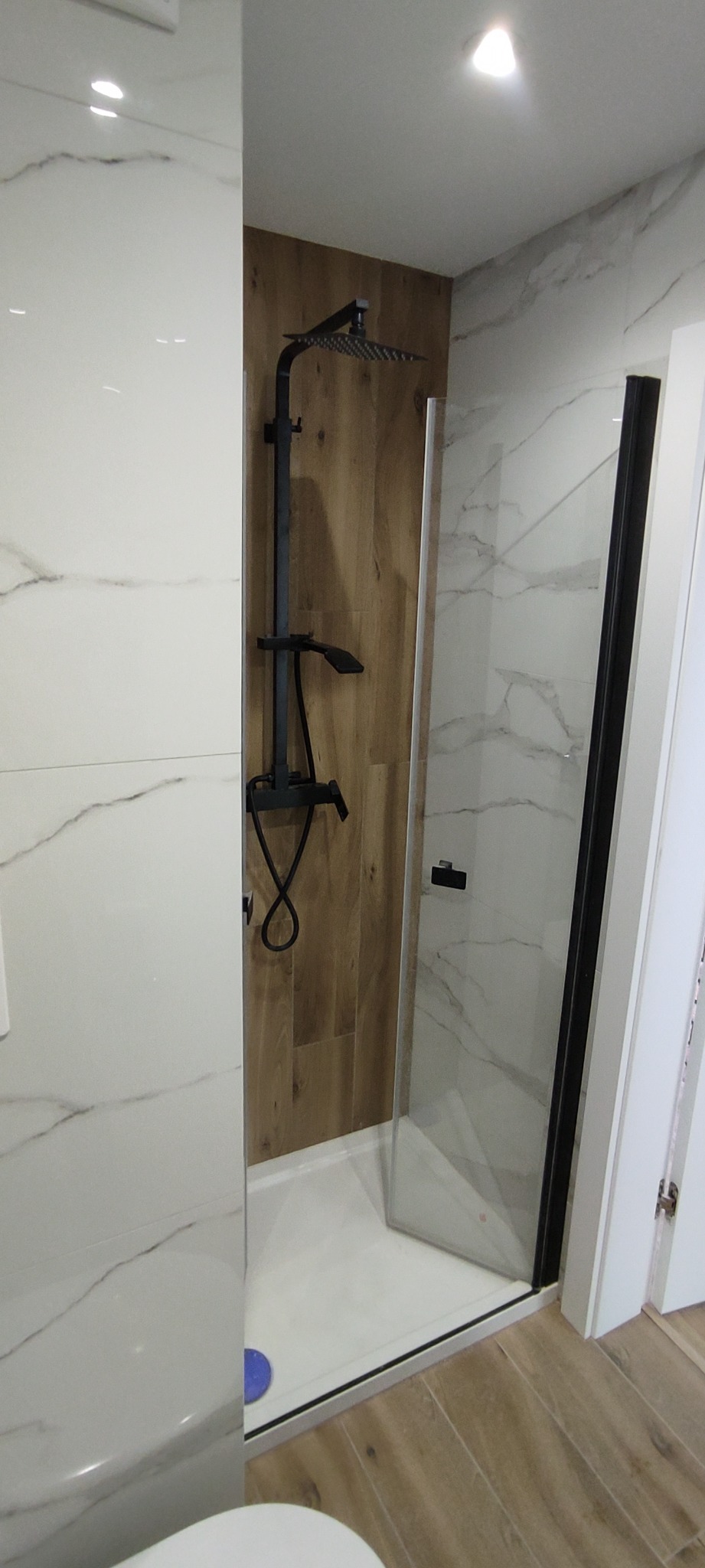 Nowoczesna kabina prysznicowa typu walk-in z czarną armaturą i panelami imitującymi drewno, wykończona białymi płytkami z marmurkowym wzorem.