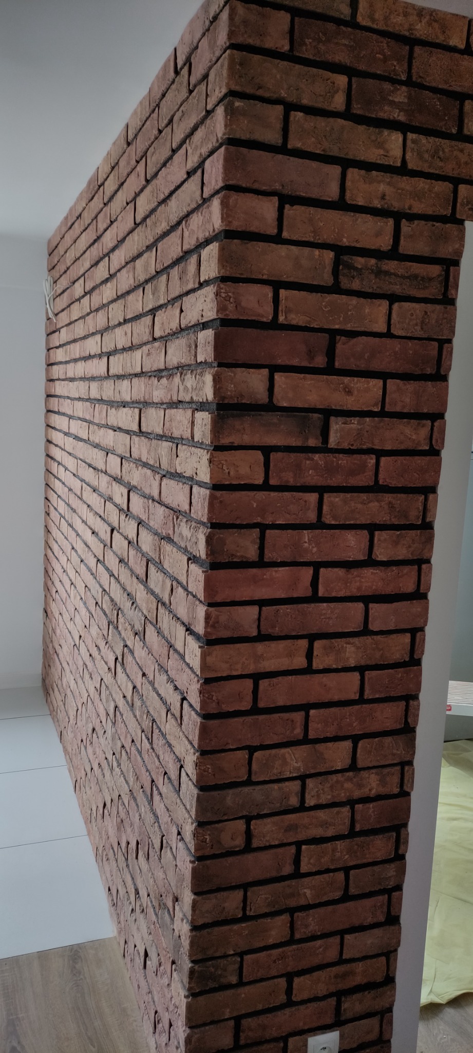 Wykończony narożnik ściany z dekoracyjnej cegły w odcieniach brązu z ciemną fugą, widoczne fragmenty podłogi z płytek i paneli oraz biała ściana w tle.