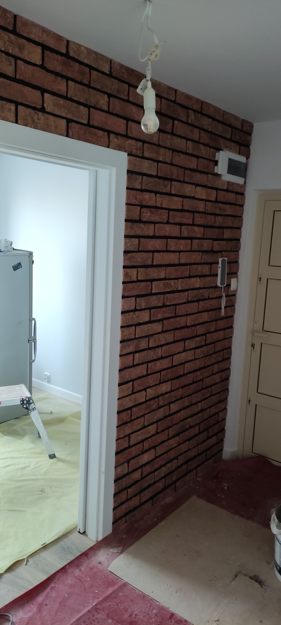 Ściana wykończona czerwoną cegłą dekoracyjną w mieszkaniu w Łomży, widoczna instalacja elektryczna, zabezpieczona podłoga, drzwi wejściowe i fragment lodówki z drabinką.