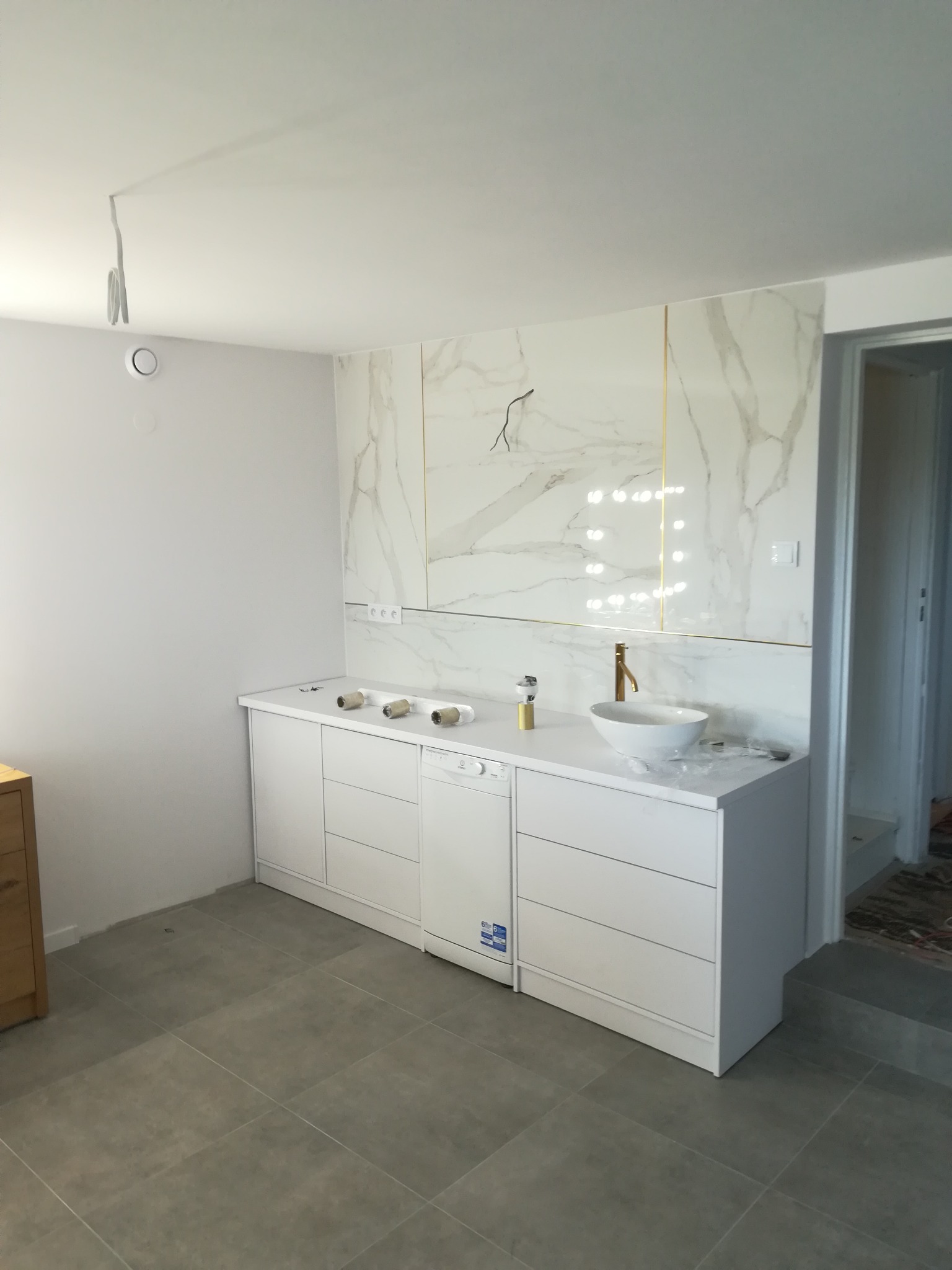Nowoczesna łazienka w trakcie wykańczania: białe szafki z umywalką nablatową i złotą armaturą, marmurowe płytki na ścianie z delikatnymi złotymi akcentami, widoczne puszki po taśmie malarskiej...