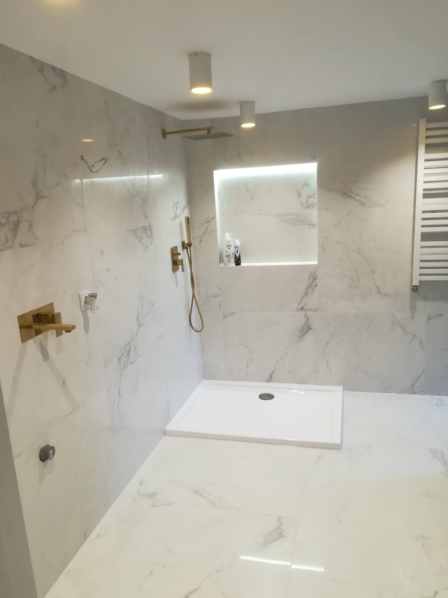 Nowoczesna łazienka wykończona białym marmurem z akcentami w kolorze złotym: armatura, deszczownica i oświetlenie w niszy ściennej.