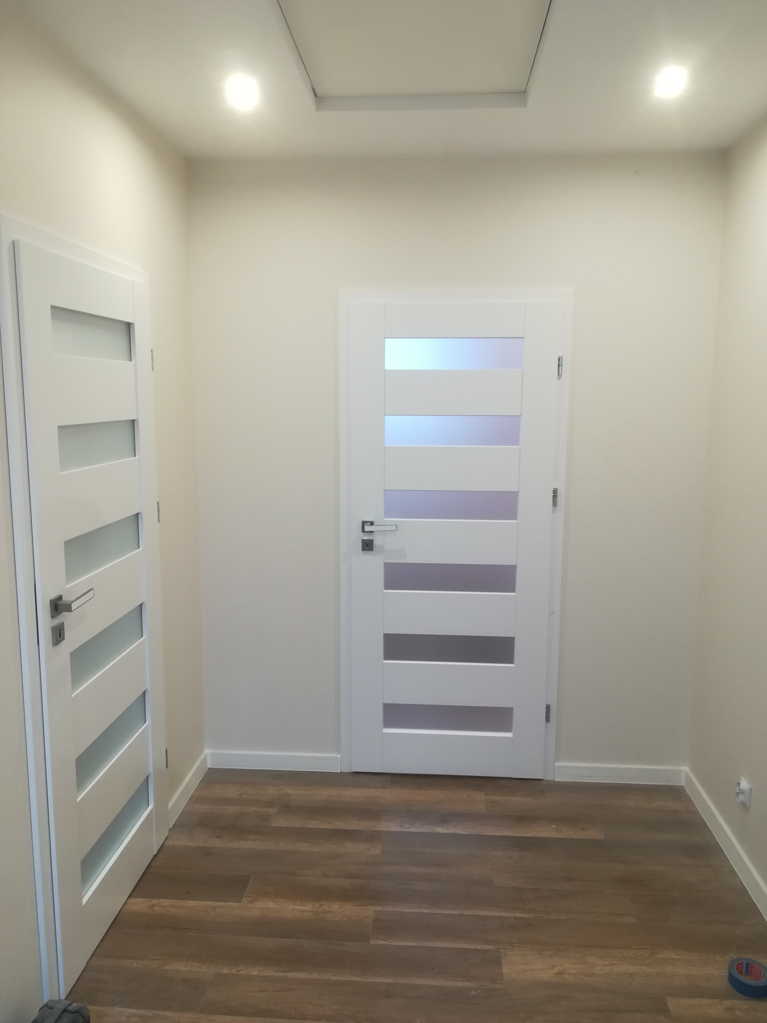 Nowoczesny przedpokój z dwiema białymi drzwiami z przeszkleniami, brązową podłogą imitującą drewno i białymi ścianami, oświetlony punktowo.