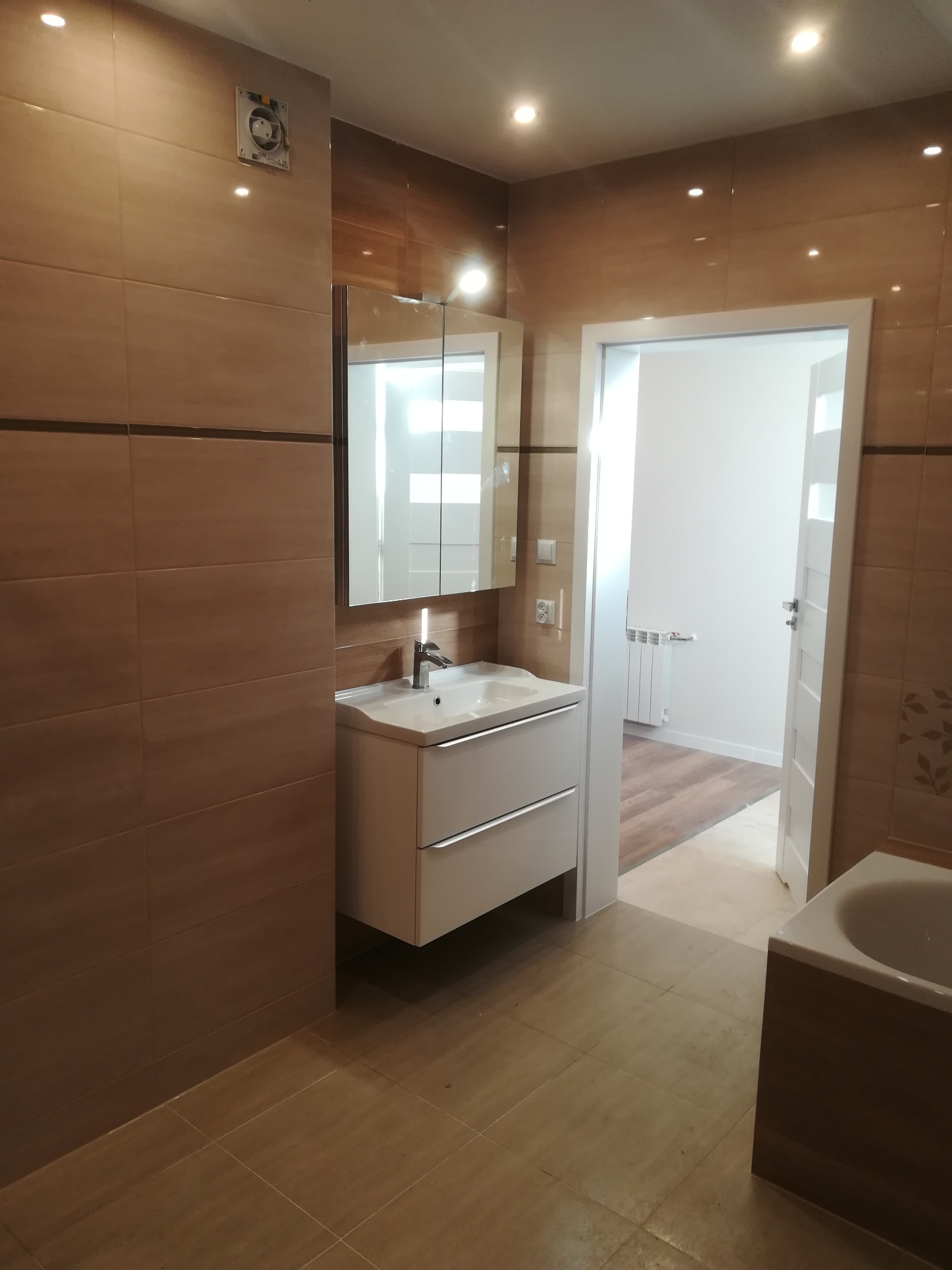 Jasna łazienka z beżowymi płytkami, umywalką z szafką i lustrem, widok na sąsiednie pomieszczenie z drewnianą podłogą i białym grzejnikiem przez otwarte drzwi.