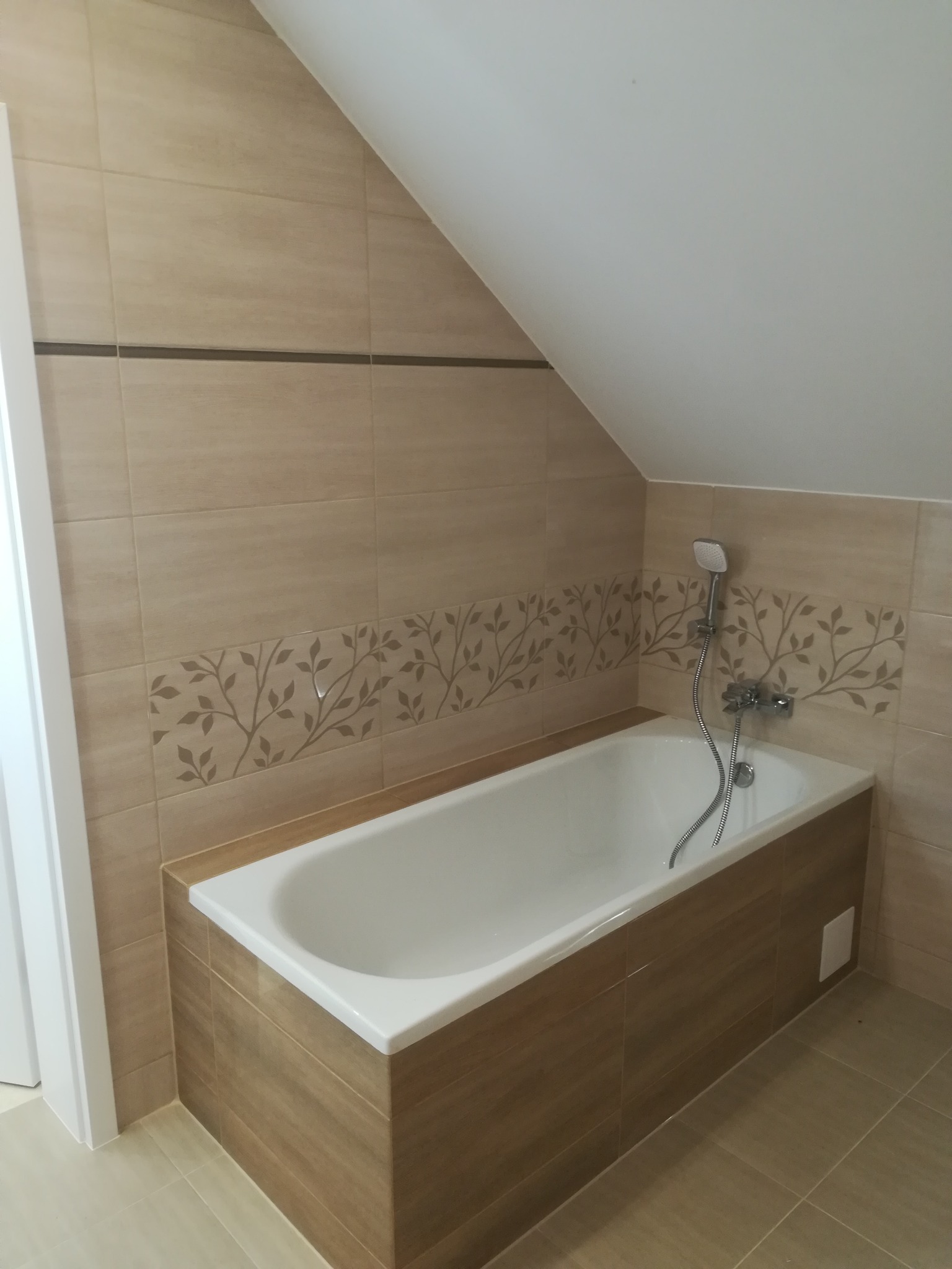 Biała wanna obudowana płytkami imitującymi drewno, z baterią i słuchawką prysznicową, na tle beżowych płytek z motywem roślinnym w łazience na poddaszu.