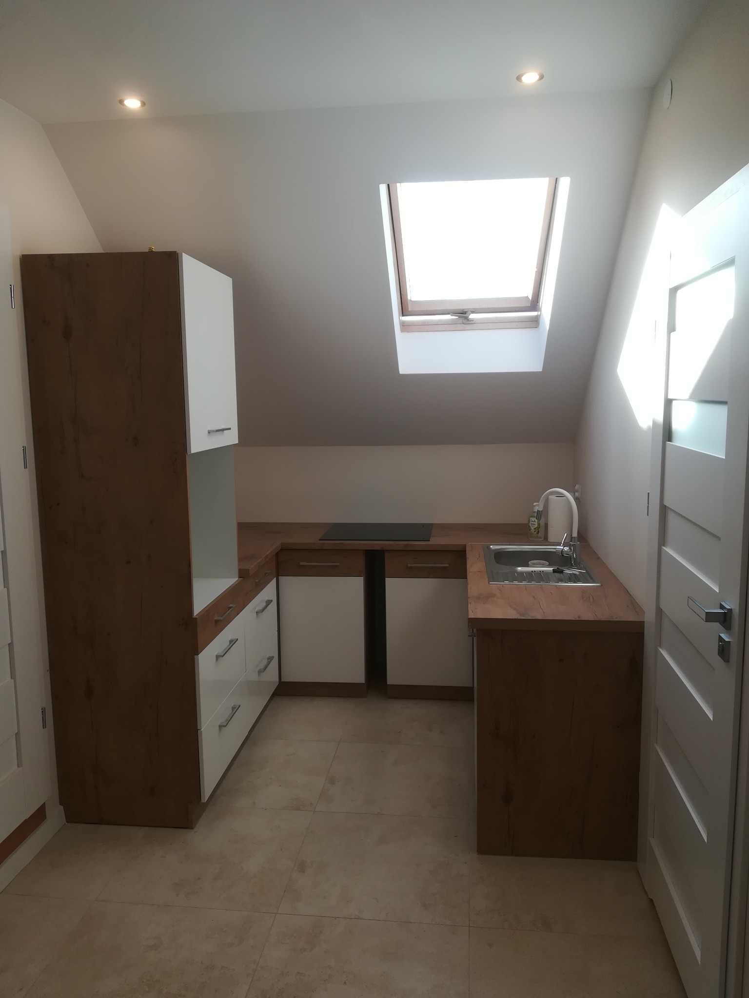 Nowoczesna kuchnia na poddaszu z drewnianymi i białymi szafkami, blatem imitującym drewno i oknem dachowym wpuszczającym naturalne światło.