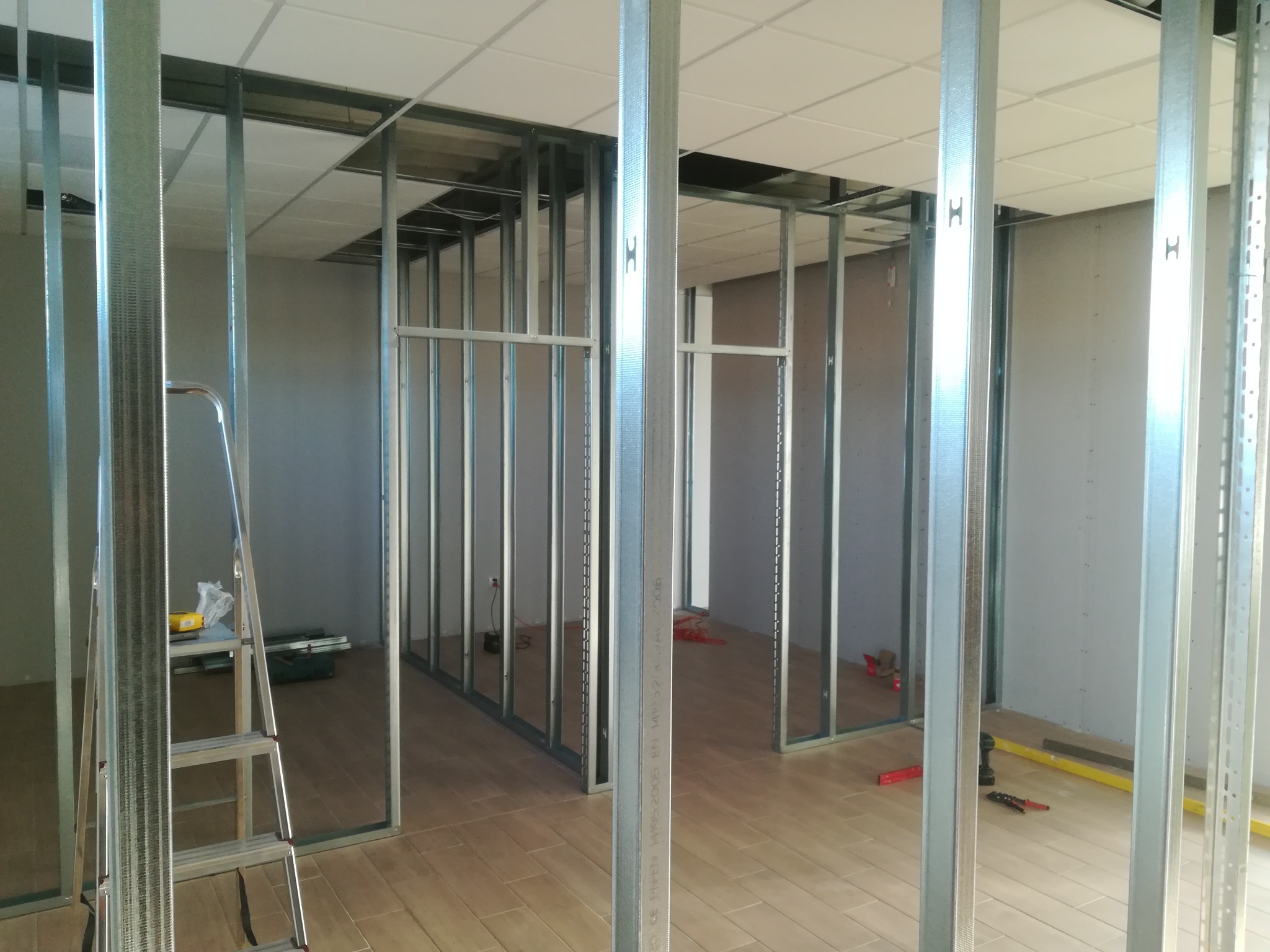Metalowa konstrukcja ścian działowych z płyt kartonowo-gipsowych w trakcie montażu, widoczna drabina i narzędzia na podłodze z płytek imitujących drewno.