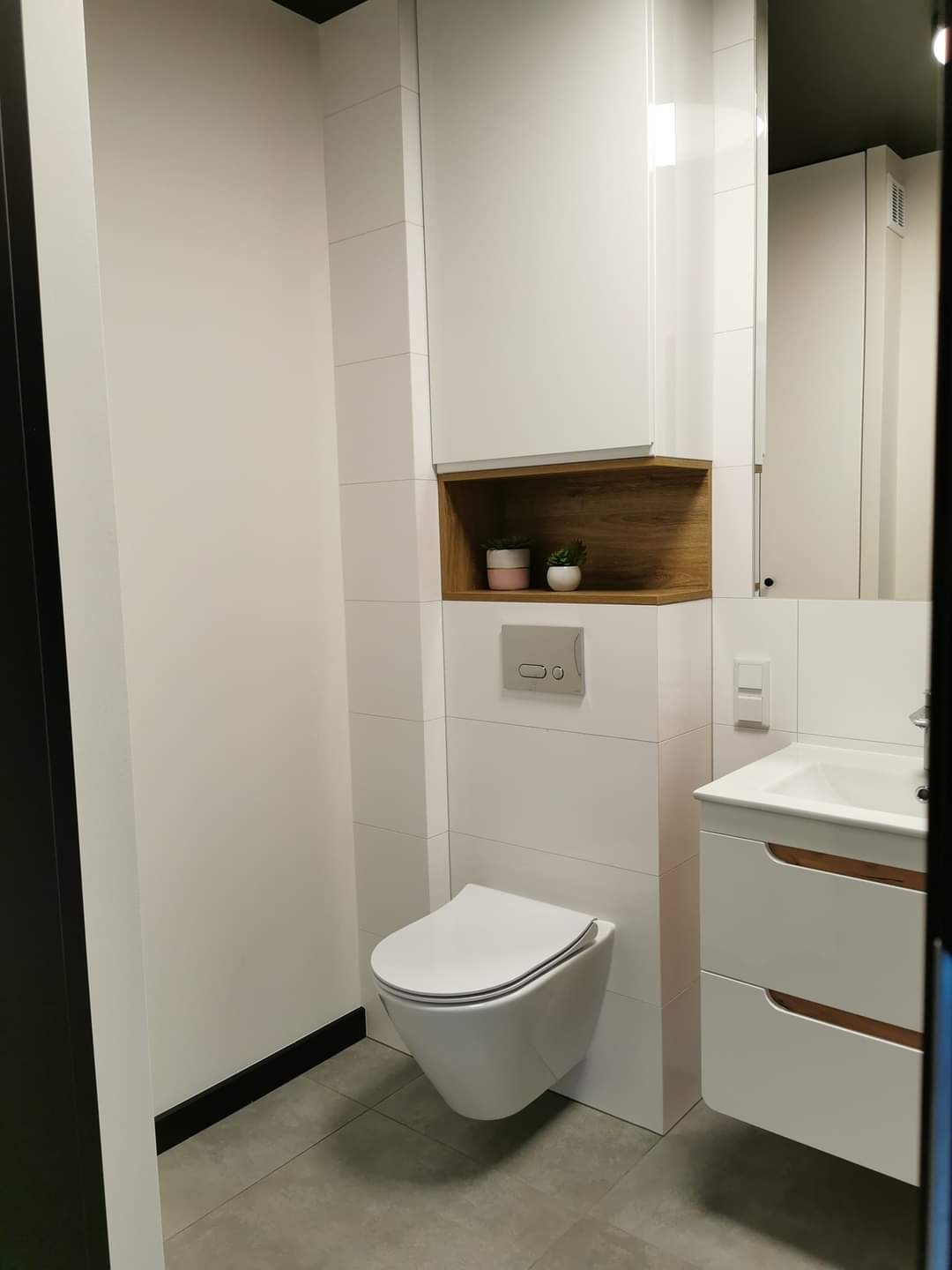 Nowoczesna łazienka z podwieszaną toaletą, umywalką z szafką z drewnianymi akcentami i wnęką z drewnianym wykończeniem, w której znajdują się dwie małe rośliny doniczkowe.