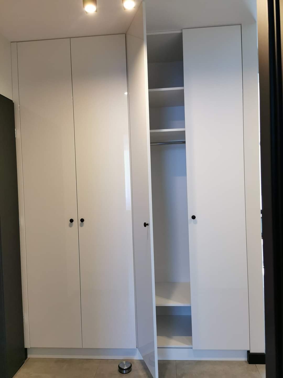 Biała, wysoka szafa z połyskiem, z czarnymi okrągłymi uchwytami, częściowo otwarta, ukazująca półki i drążek na ubrania wewnątrz, z widocznym elementem podłogowym w dolnym rogu.