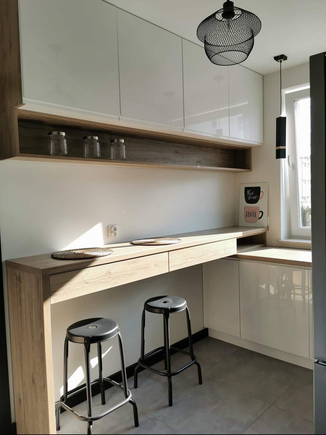 Nowoczesna kuchnia z białymi, błyszczącymi szafkami górnymi, blatem z drewna w jasnym odcieniu, dwoma czarnymi hokerami i dekoracyjnym oświetleniem w postaci ażurowej lampy.