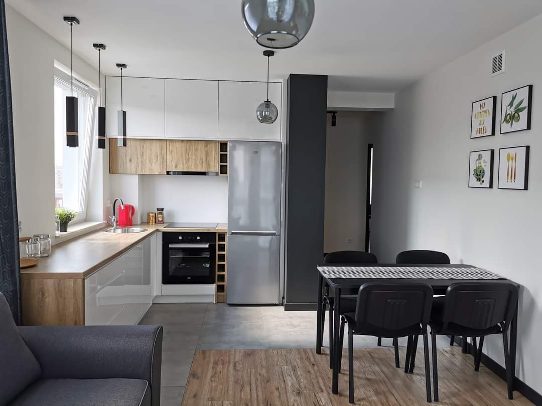 Nowoczesna kuchnia z białymi szafkami górnymi i drewnopodobnymi dolnymi, srebrna lodówka, czarny stół z krzesłami, oświetlenie punktowe i wiszące lampy, dekoracje ścienne.