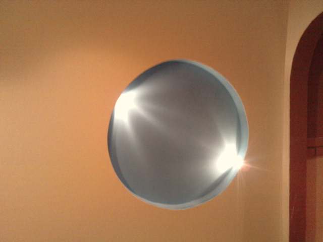 Okrągły otwór w pomarańczowej ścianie, oświetlony od wewnątrz, z fragmentem łuku architektonicznego po prawej stronie.