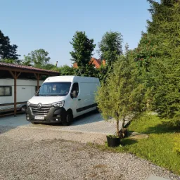Biały bus Renault Master zaparkowany na żwirowym podjeździe obok przyczepy kempingowej i iglaków w słoneczny dzień.