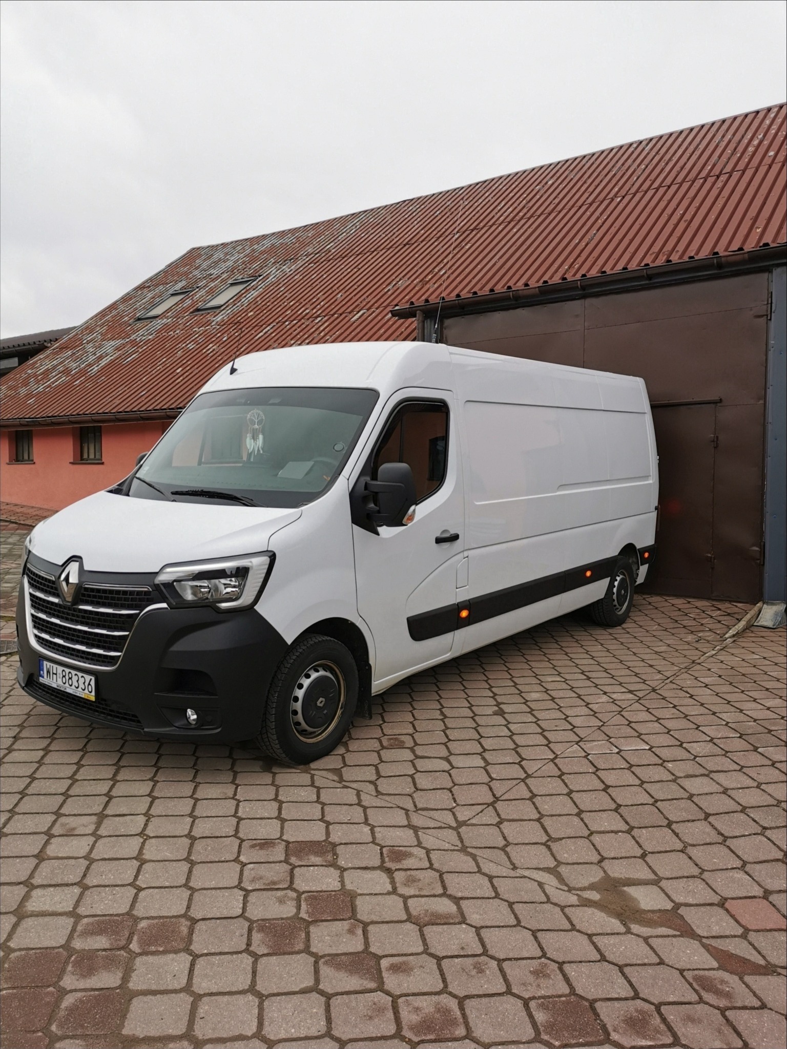 Biały Renault Master zaparkowany na brukowanym podjeździe przed garażem z brązowymi drzwiami i budynkiem z czerwoną dachówką w tle. Na przedniej szybie widoczny łapacz snów.