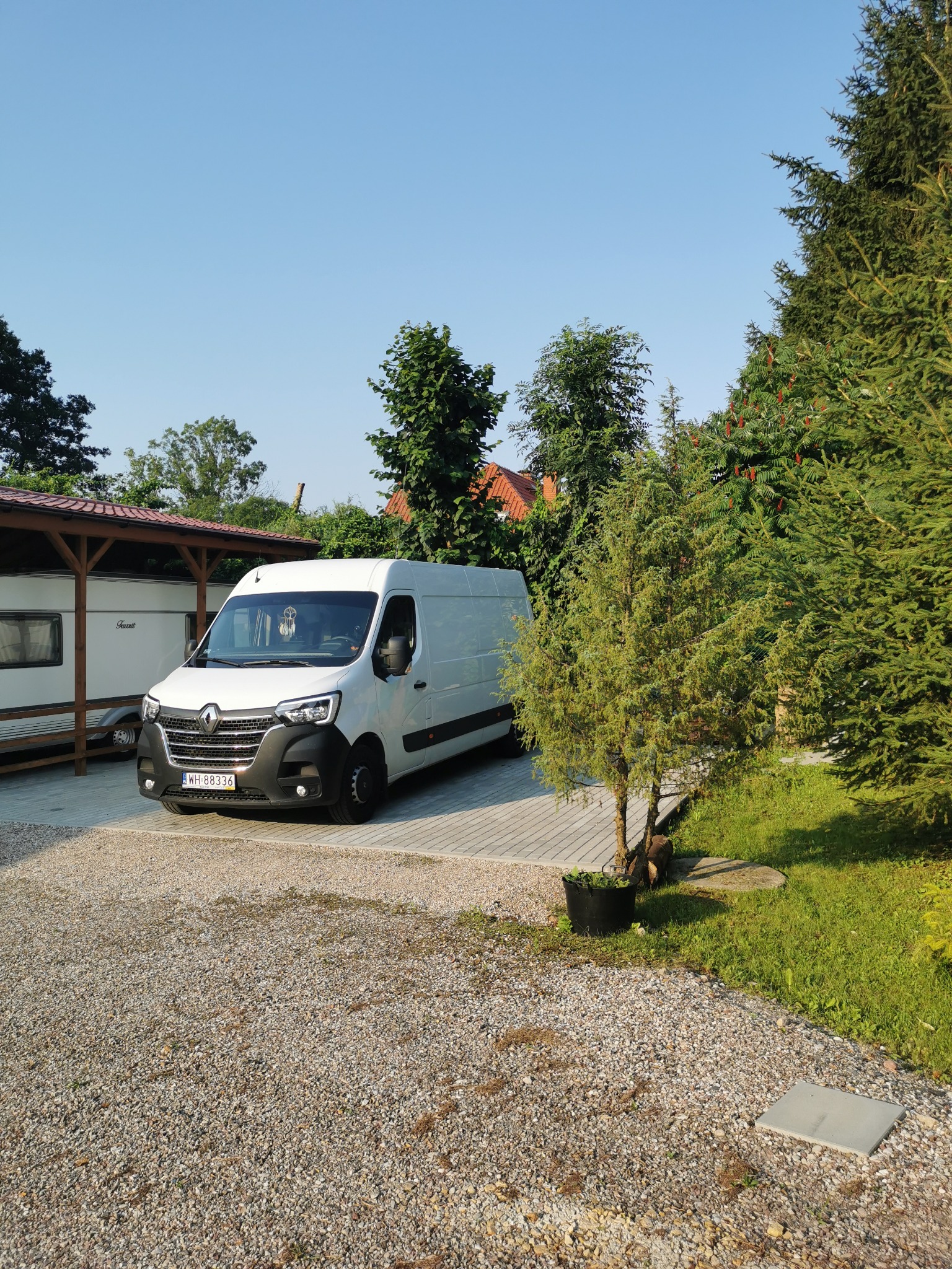 Biały bus Renault Master zaparkowany na żwirowym podjeździe obok przyczepy kempingowej i iglaków w słoneczny dzień.
