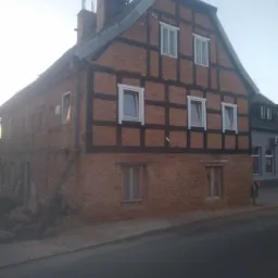 Dwukondygnacyjny budynek z muru pruskiego z ceglaną elewacją, widoczne ślady renowacji i przygotowania do prac wykończeniowych, w tle fragment ulicy.