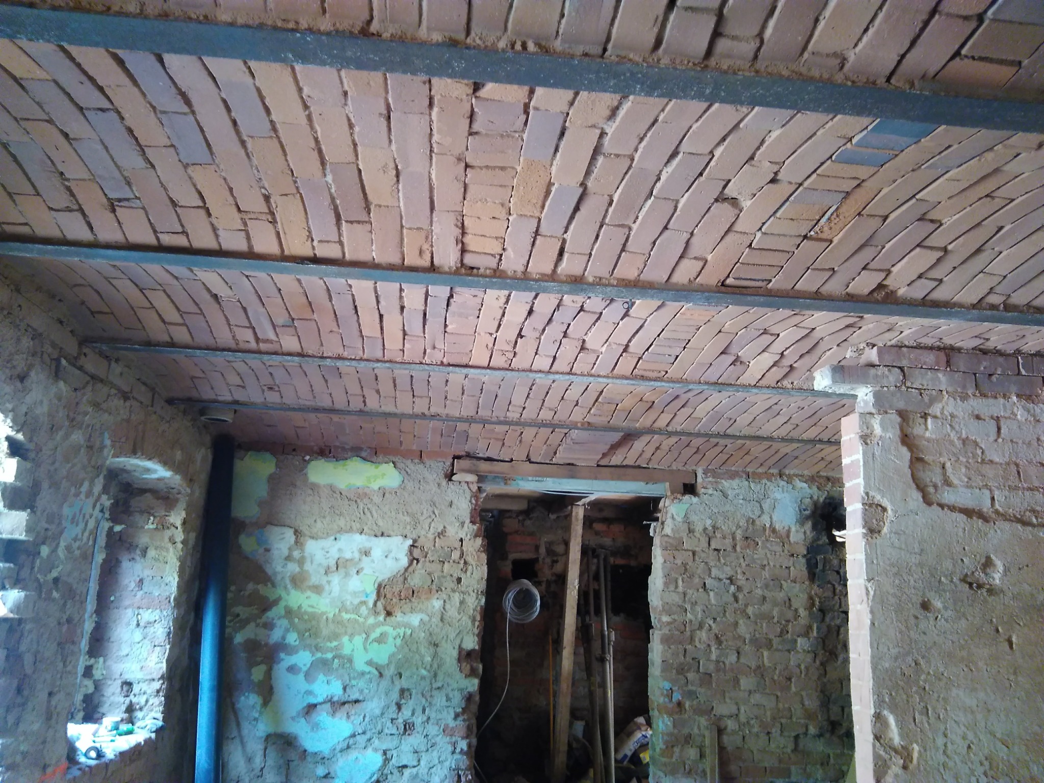 Wnętrze remontowanego pomieszczenia z ceglanym sklepieniem łukowym podpartym metalowymi belkami; ściany z widocznymi śladami starego tynku i cegieł, otwór drzwiowy z widocznymi przewodami...