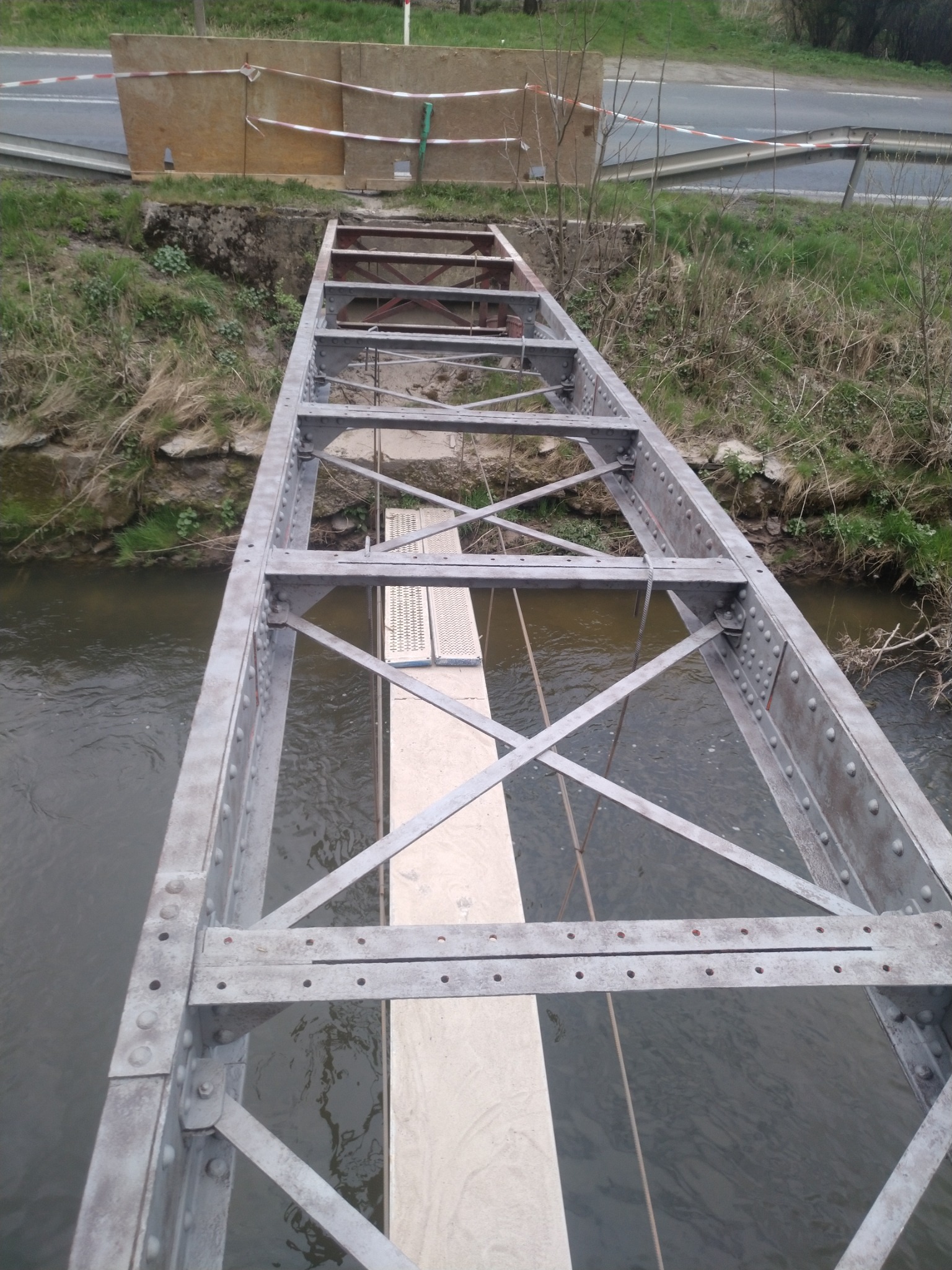 Metalowa konstrukcja mostu nad rzeką, widok z góry, z elementami drewnianymi i zabezpieczeniami budowlanymi w tle.