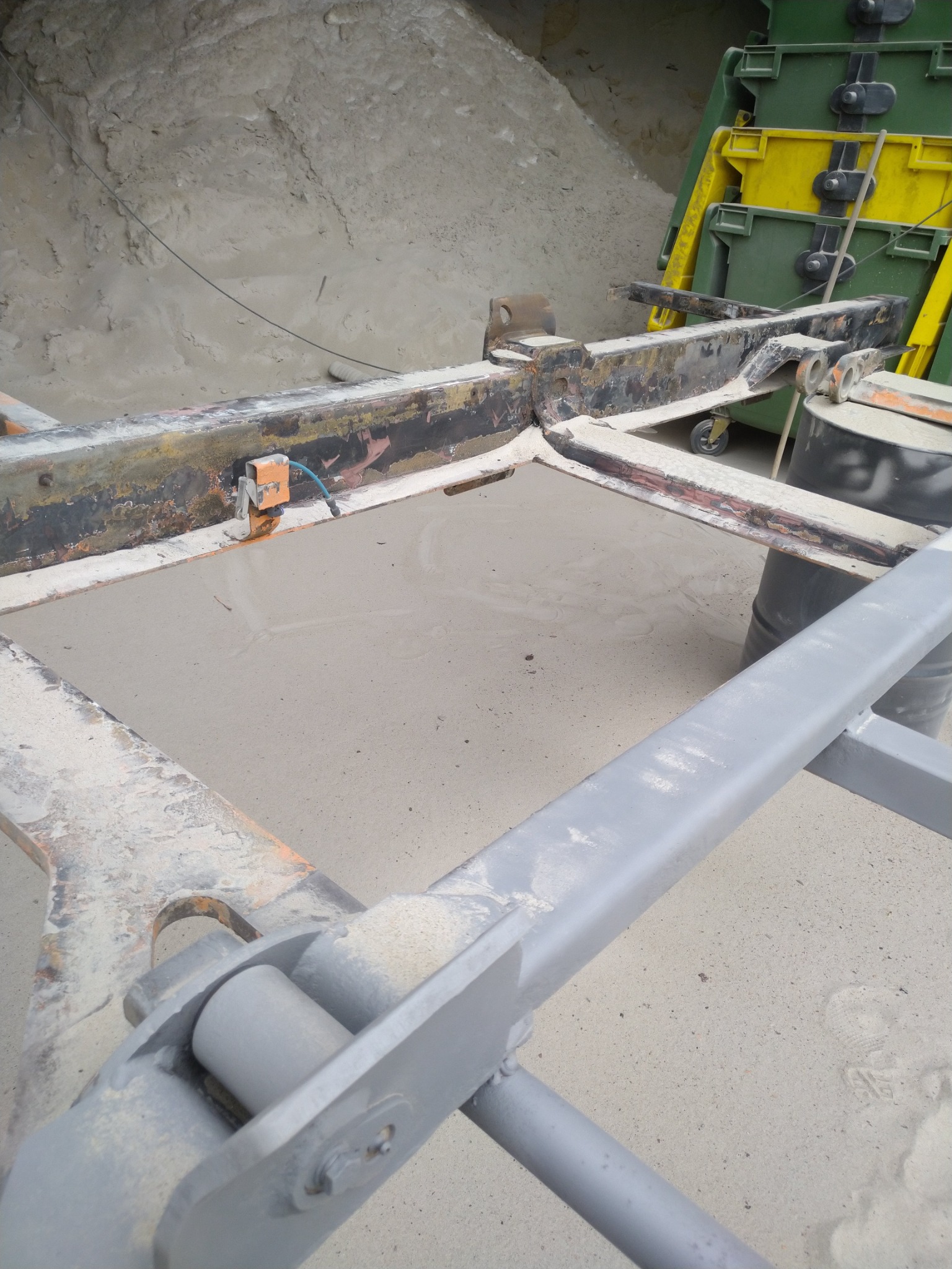 Metalowa konstrukcja z pozostałościami rdzy i śladami obróbki, wypełniona szarym materiałem przypominającym piasek, na tle hałdy tego samego materiału i plastikowych pojemników.