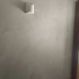 Ściana w pokoju z dekoracyjną szarą gładzią wenecką i białym włącznikiem światła, widoczna listwa przypodłogowa i fragment ościeżnicy drzwi.