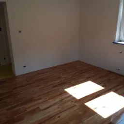 Wykończony pokój z panelami podłogowymi w odcieniach brązu, wpadające promienie słoneczne podkreślają połysk podłogi, ściany pomalowane na jasno, widoczne gniazdka elektryczne i fragment folii...