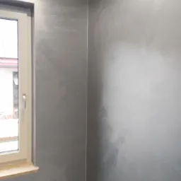 Ściana w rogu pokoju pokryta dekoracyjną szarą farbą o efekcie chmur, widoczne okno z jasną ramą i parapetem.