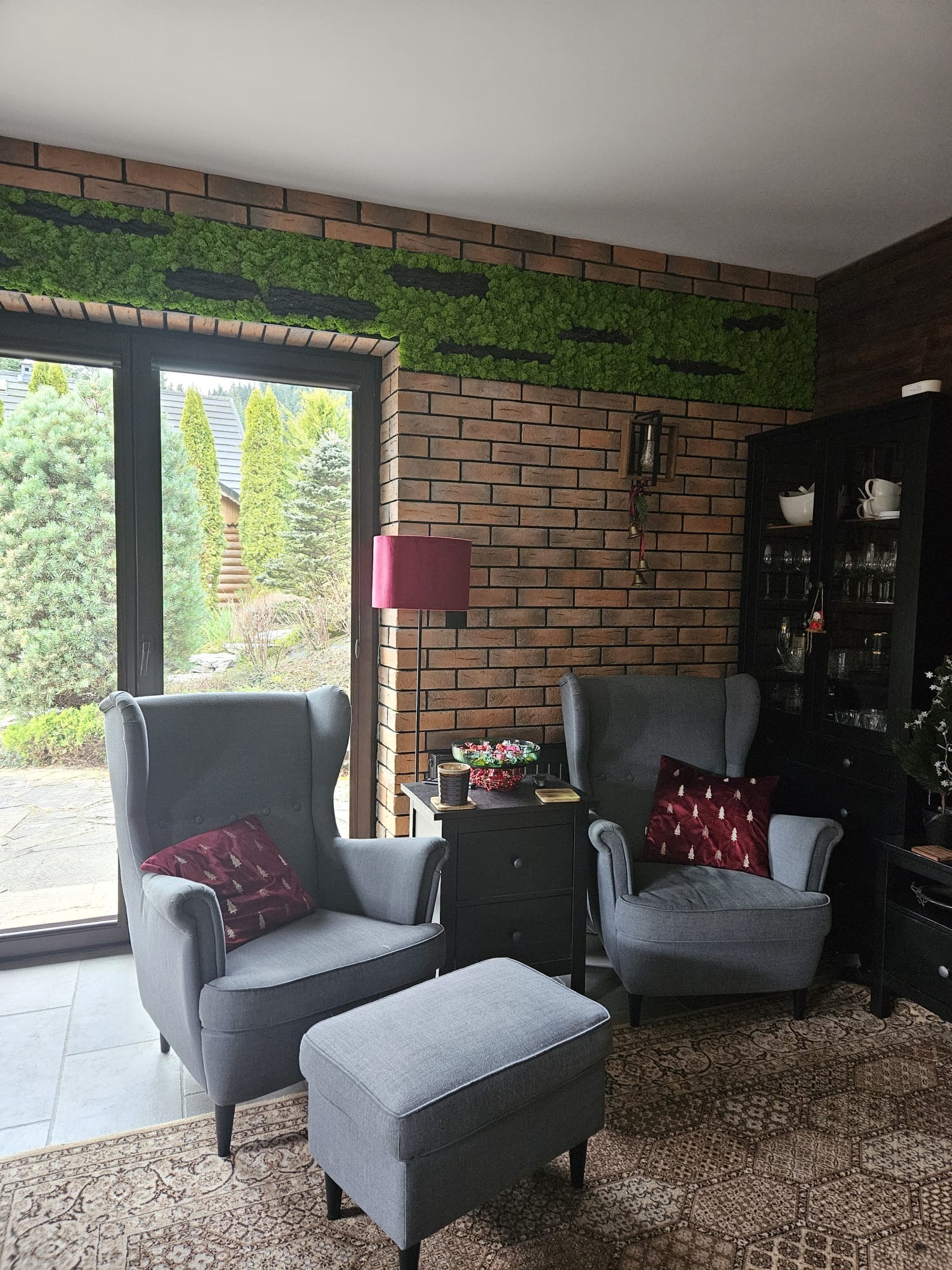 Salon z ceglaną ścianą, dekoracją z mchu, szarymi fotelami typu uszak i dywanem w stylu vintage.