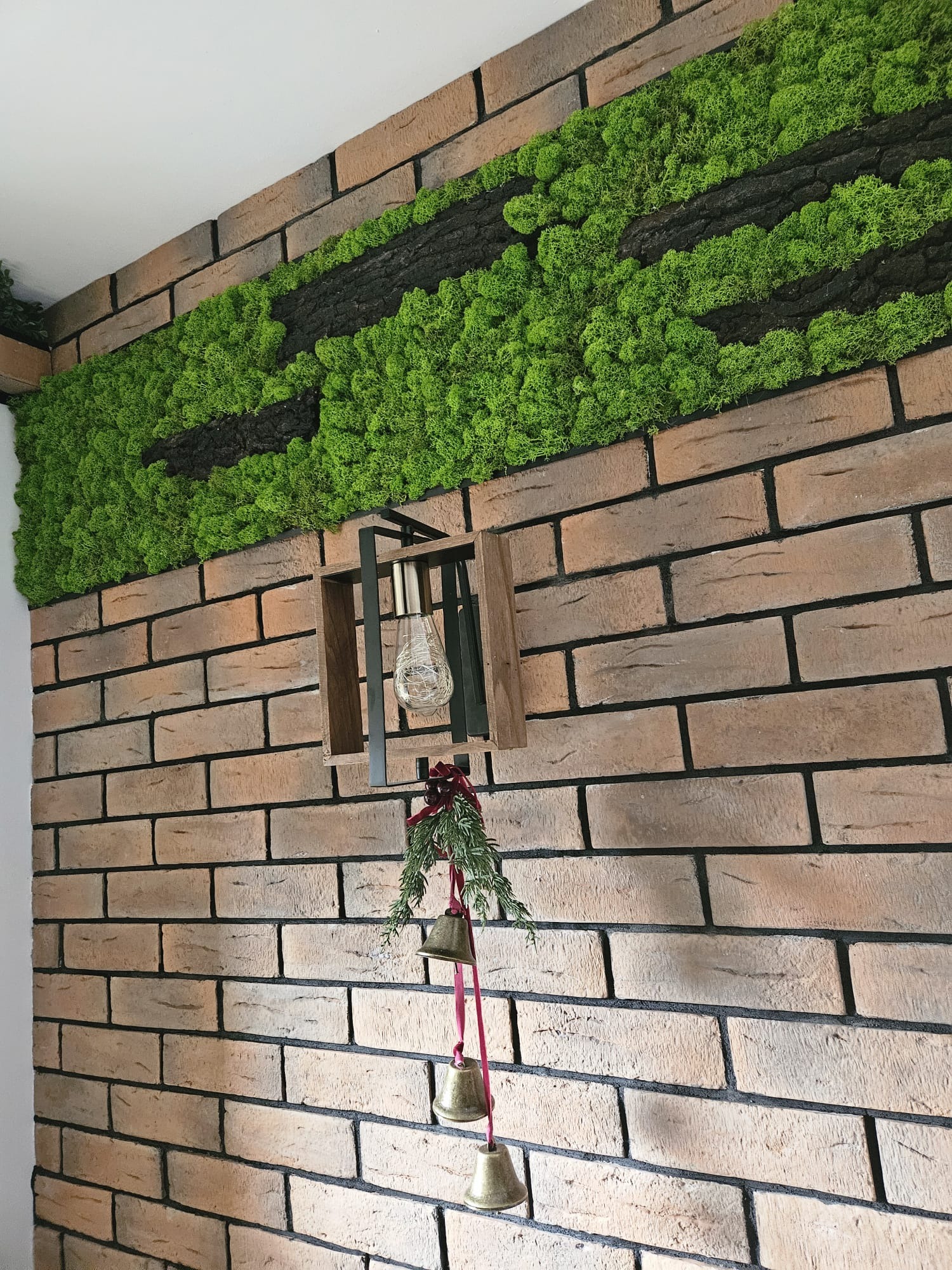 Ściana z cegły dekoracyjnej z paskiem mchu i kory, ozdobiona wiszącą lampą w drewnianej obudowie i świątecznymi dzwoneczkami na czerwonej wstążce.