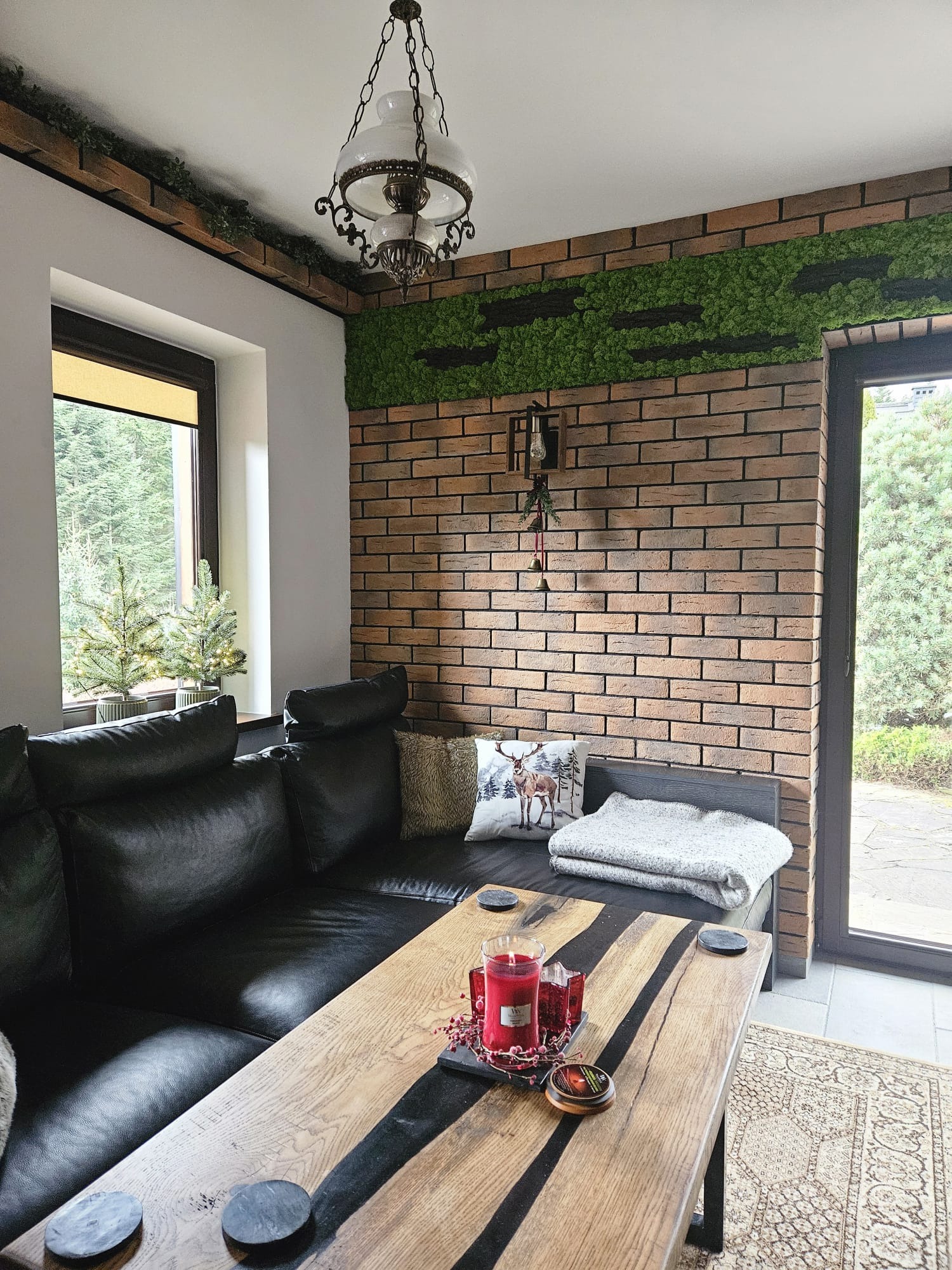 Salon z ceglaną ścianą ozdobioną mchem, czarną skórzaną sofą i drewnianym stolikiem kawowym z żywicą epoksydową; dekoracje świąteczne i widok na zewnątrz przez okno.