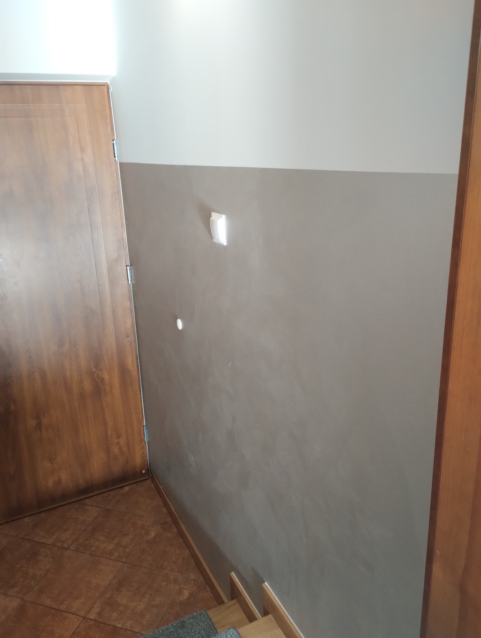 Ściana pomalowana na dwa kolory, z widocznym włącznikiem światła i otworem po kołku, brązowe płytki na podłodze, drewniane drzwi po lewej stronie