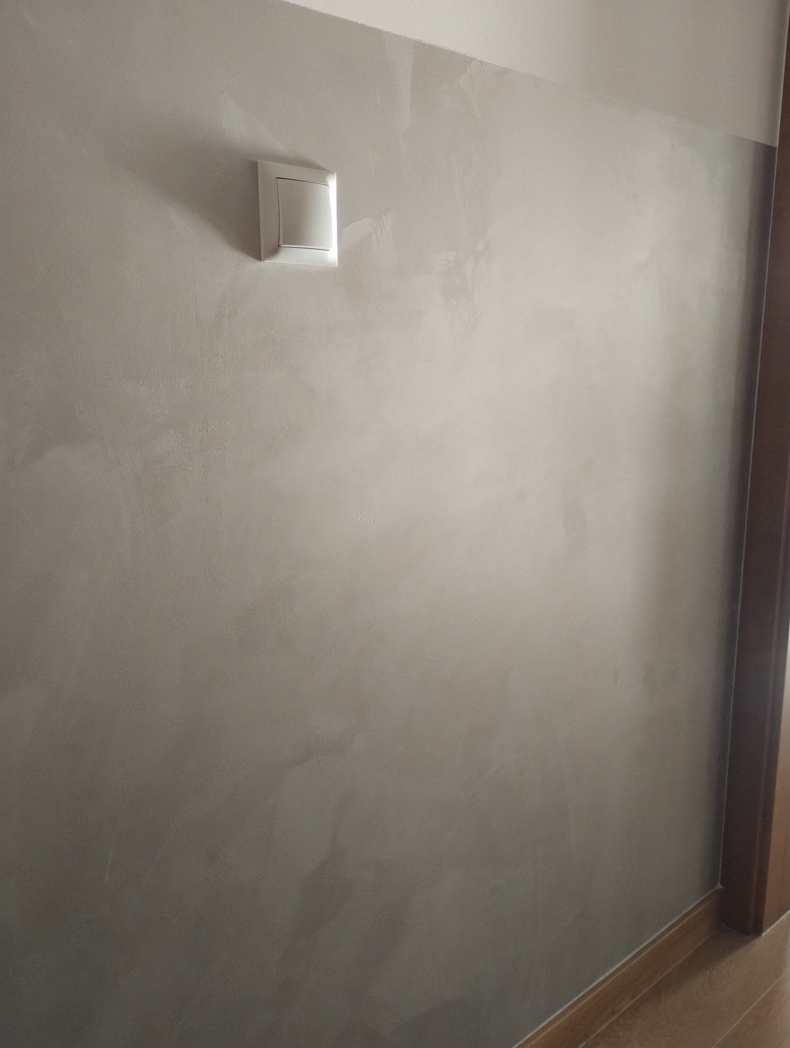 Ściana w pokoju z dekoracyjną szarą gładzią wenecką i białym włącznikiem światła, widoczna listwa przypodłogowa i fragment ościeżnicy drzwi.
