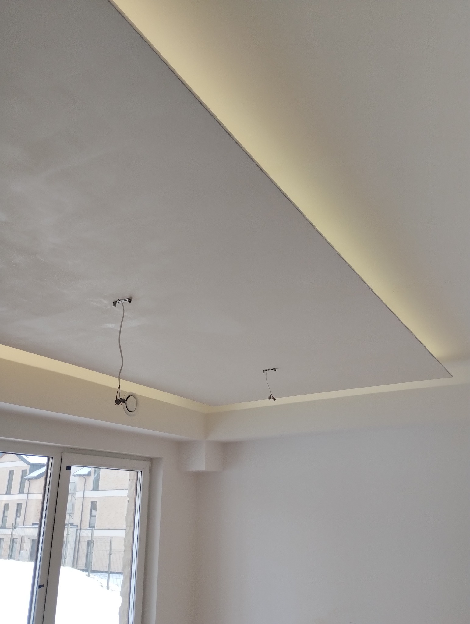 Płyta karton gips na suficie z wbudowanym oświetleniem LED, widoczne kable od oświetlenia, okno z widokiem na zaśnieżone budynki.