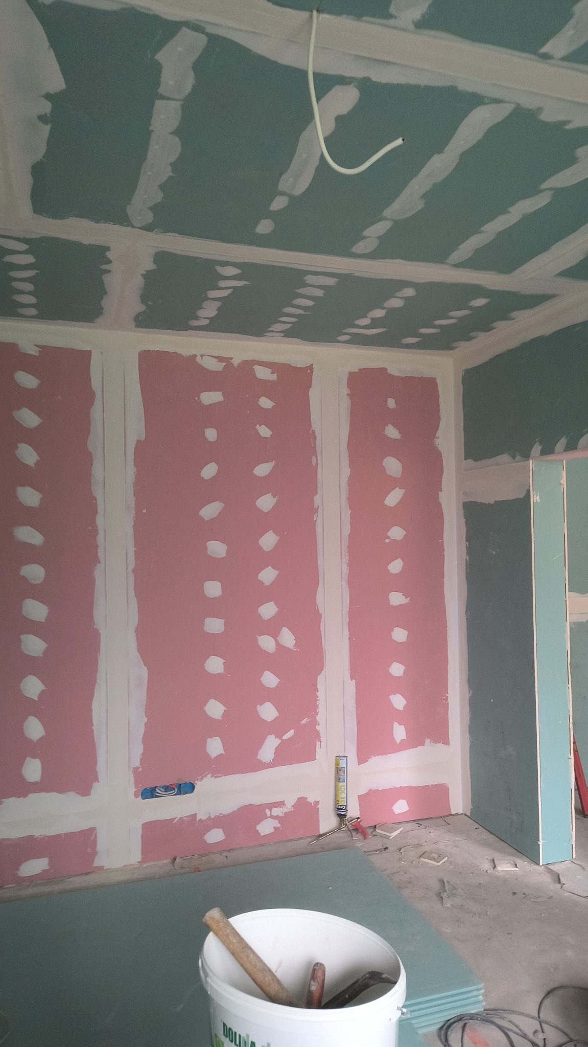 Wnętrze w trakcie montażu płyt kartonowo-gipsowych: ściany z widocznymi łączeniami, sufit z instalacją elektryczną, wiadro z narzędziami na podłodze.