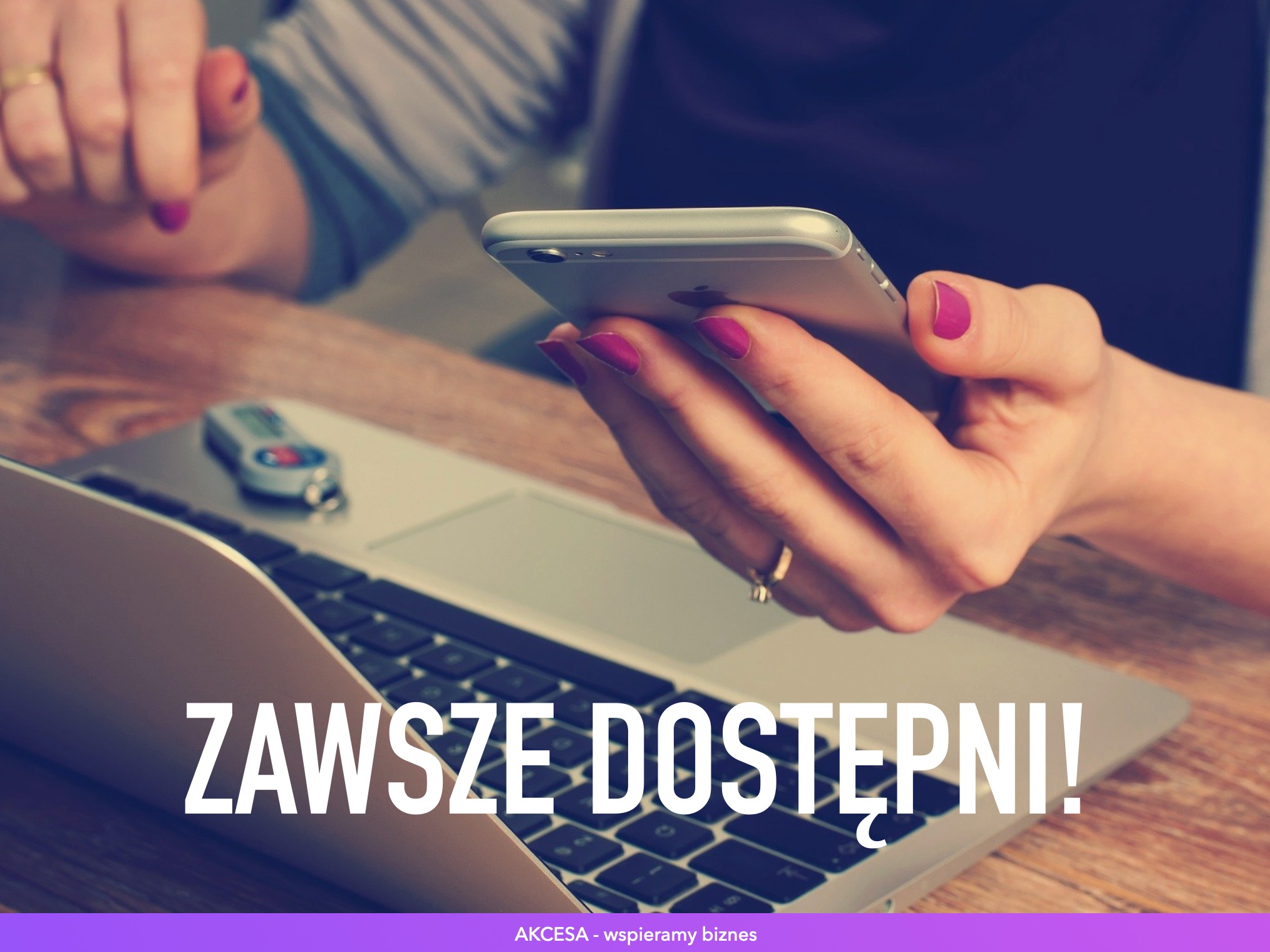 Dłoń z fuksjowym lakierem trzyma smartfon nad otwartym laptopem na drewnianym biurku, obok kalkulator, napis 'Zawsze dostępni!'