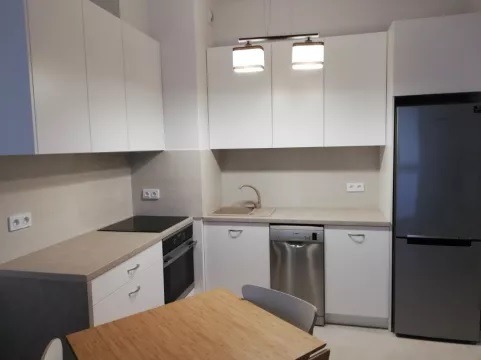 Jasna kuchnia z białymi szafkami, szarym blatem i srebrną lodówką, oświetlona lampą sufitową z dwoma abażurami, częściowo widoczny drewniany stół.