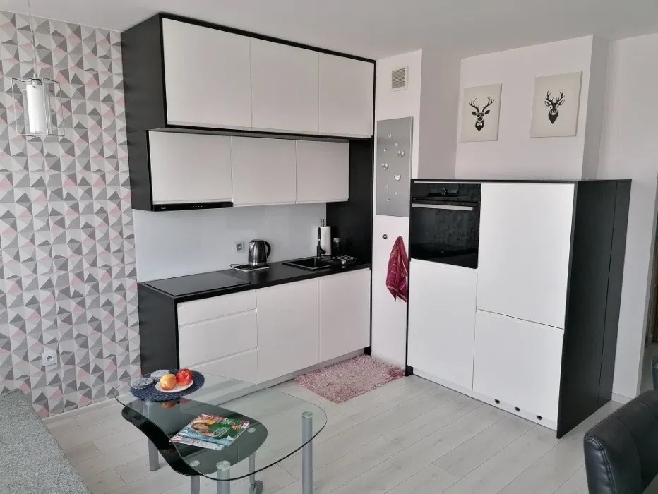 Nowoczesna, minimalistyczna kuchnia w bieli i czerni, z geometryczną tapetą i szklanym stolikiem kawowym z owocami, oświetlona wiszącą lampą.