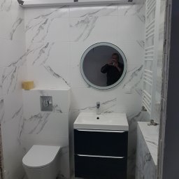 Dariusz Stokowski - Nowoczesna łazienka z białymi płytkami imitującymi marmur, okrągłe lustro z podświetleniem, wisząca toaleta, umywalka z szafką w kolorze granatowym, grzejnik drabinkowy i żółta gąbka na zabudowie...