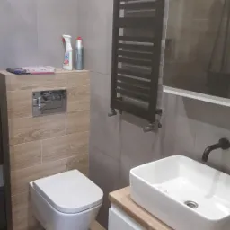 Nowoczesna łazienka z szarymi płytkami, drewnopodobnymi akcentami, wiszącą toaletą, grzejnikiem drabinkowym i umywalką nablatową na szafce z drewnianym blatem.