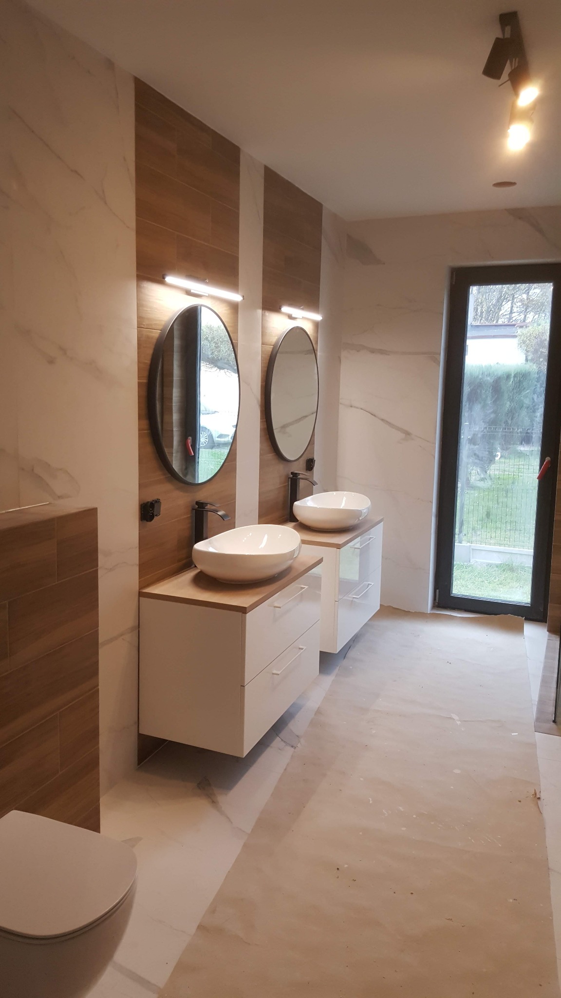 Wykończona łazienka w Kania Góra z dwoma umywalkami nablatowymi na białych szafkach, okrągłe lustra z podświetleniem, płytki imitujące drewno i marmur, oraz duże okno z widokiem na zewnątrz.