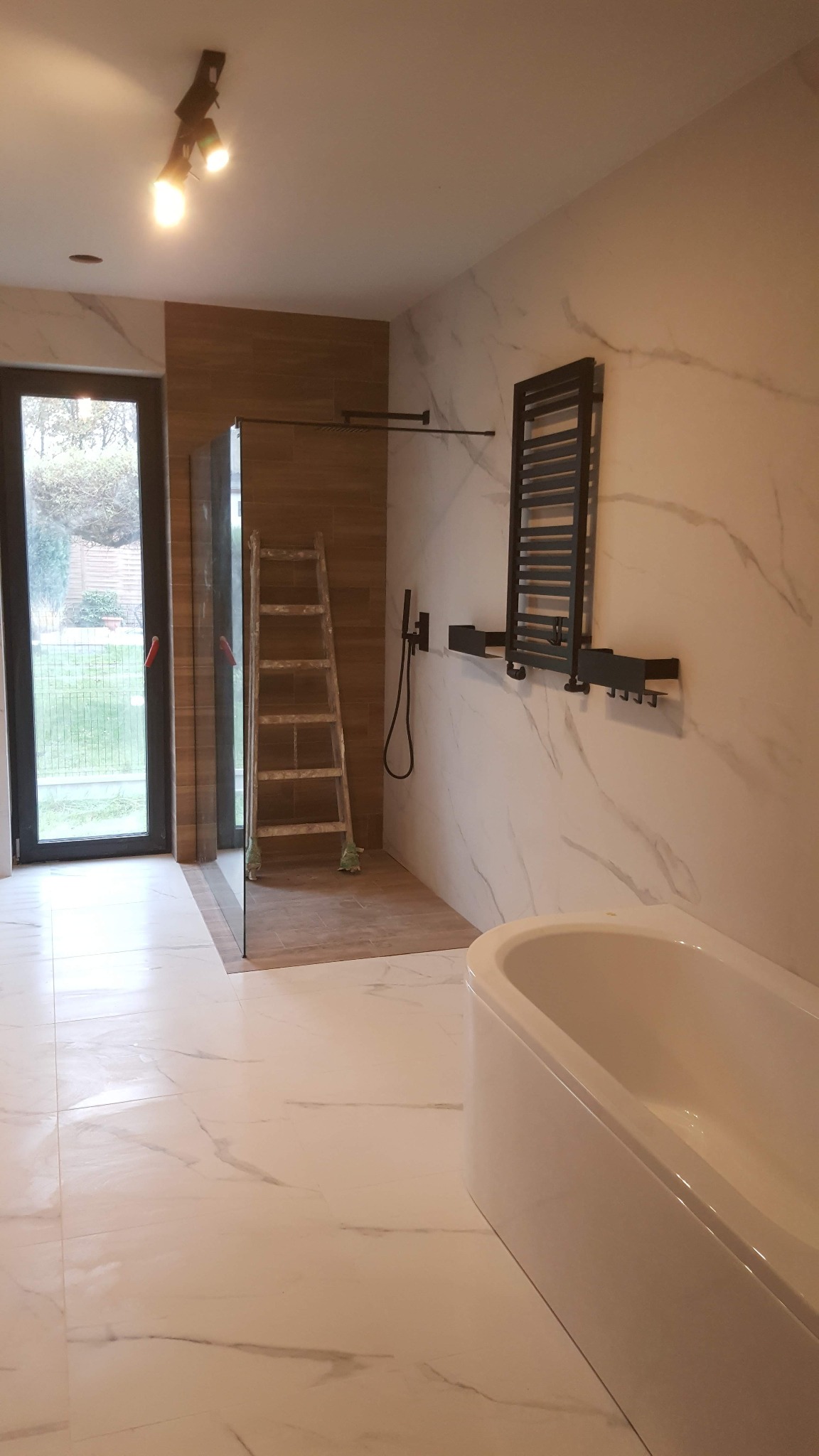 Nowoczesna łazienka w trakcie wykańczania: białe marmurowe płytki, kabina prysznicowa ze szklaną ścianką, drabina oparta o ścianę z brązowych płytek, grzejnik drabinkowy w kolorze czarnym, biała...