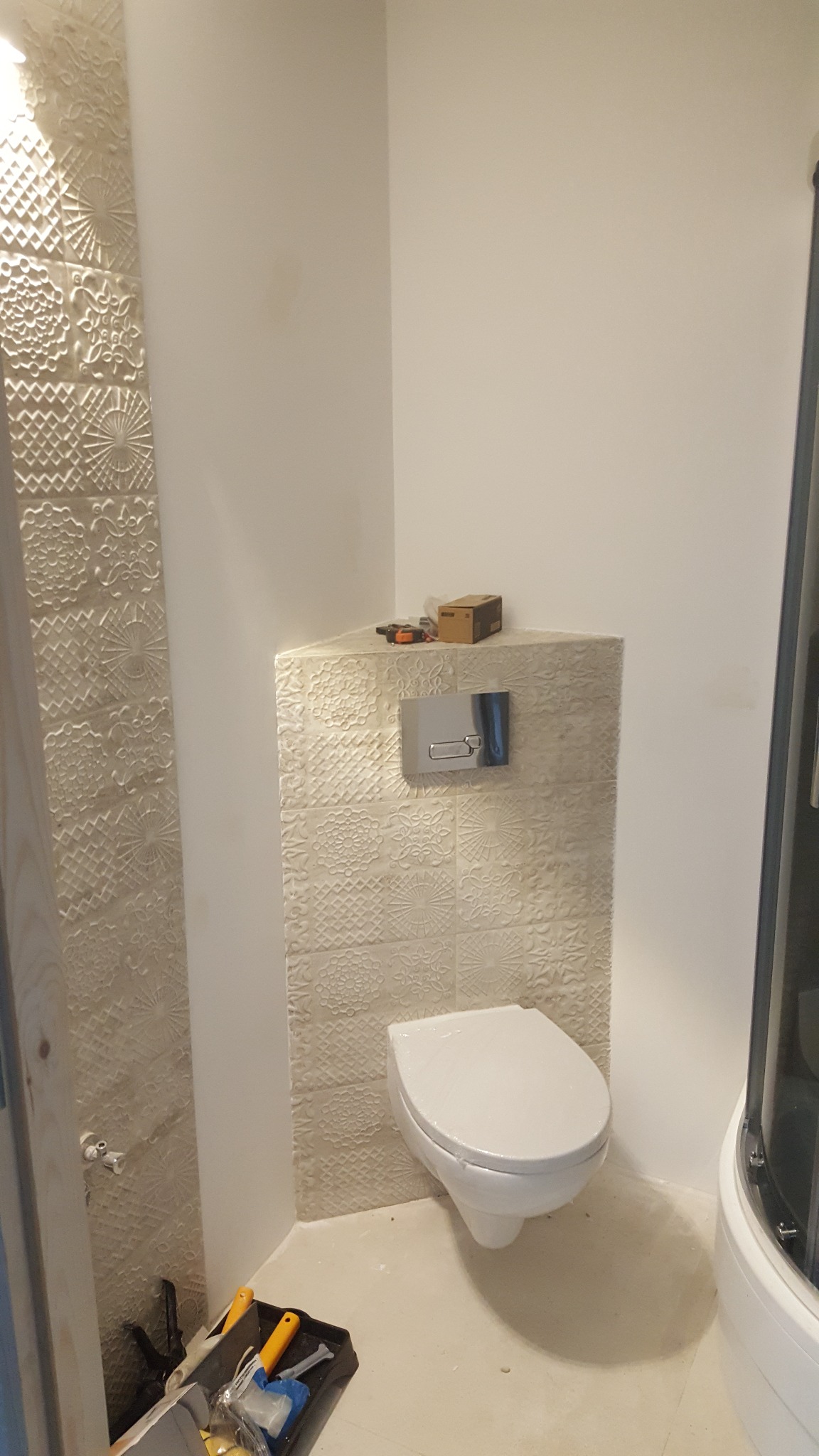 Wisząca toaleta z ukrytą spłuczką, obudowana płytkami z tłoczonym wzorem, w trakcie montażu. Na górze obudowy leżą narzędzia i pudełko, a na podłodze widoczny kuweta z narzędziami.