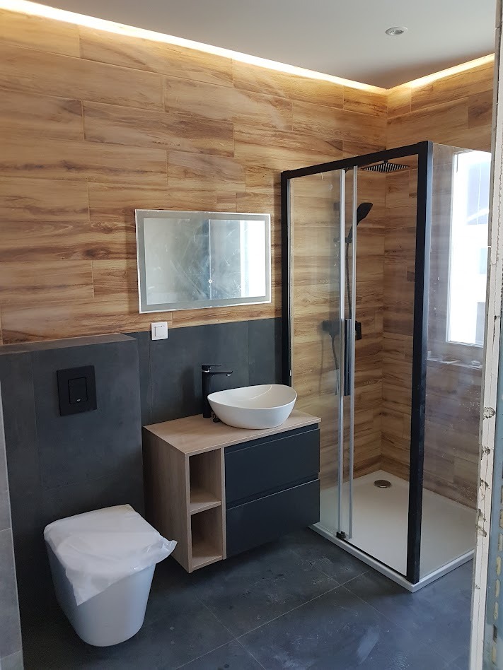 Nowoczesna łazienka z drewnopodobnymi płytkami ściennymi, kabiną prysznicową z czarną ramą, umywalką nablatową na szafce z szufladami i podświetlanym lustrem.