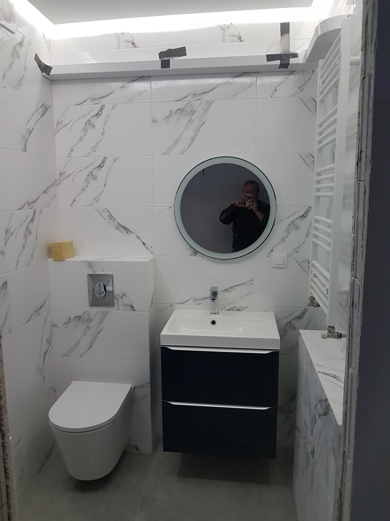 Nowoczesna łazienka z białymi płytkami imitującymi marmur, okrągłe lustro z podświetleniem, wisząca toaleta, umywalka z szafką w kolorze granatowym, grzejnik drabinkowy i żółta gąbka na zabudowie...