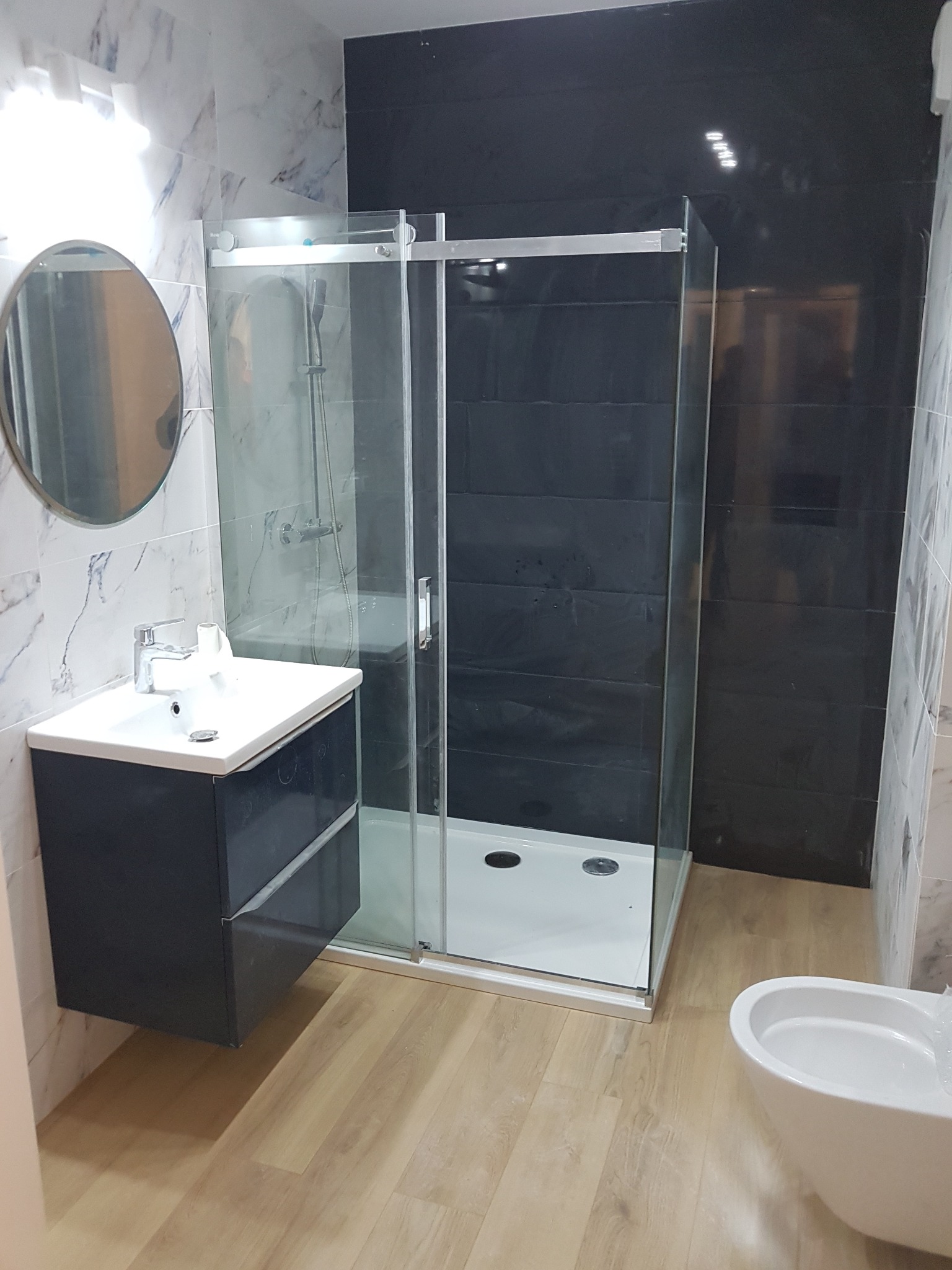Wyremontowana łazienka z kabiną prysznicową z przesuwnymi drzwiami, umywalką z szafką w kolorze granatowym i owalnym lustrem na ścianie z marmurowym wzorem.