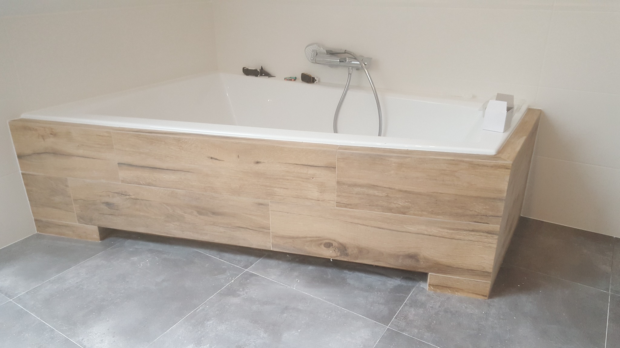 Obudowana wanna z drewnopodobnymi płytkami, szara podłoga z płytek, zabawki na wannie i białe ściany.