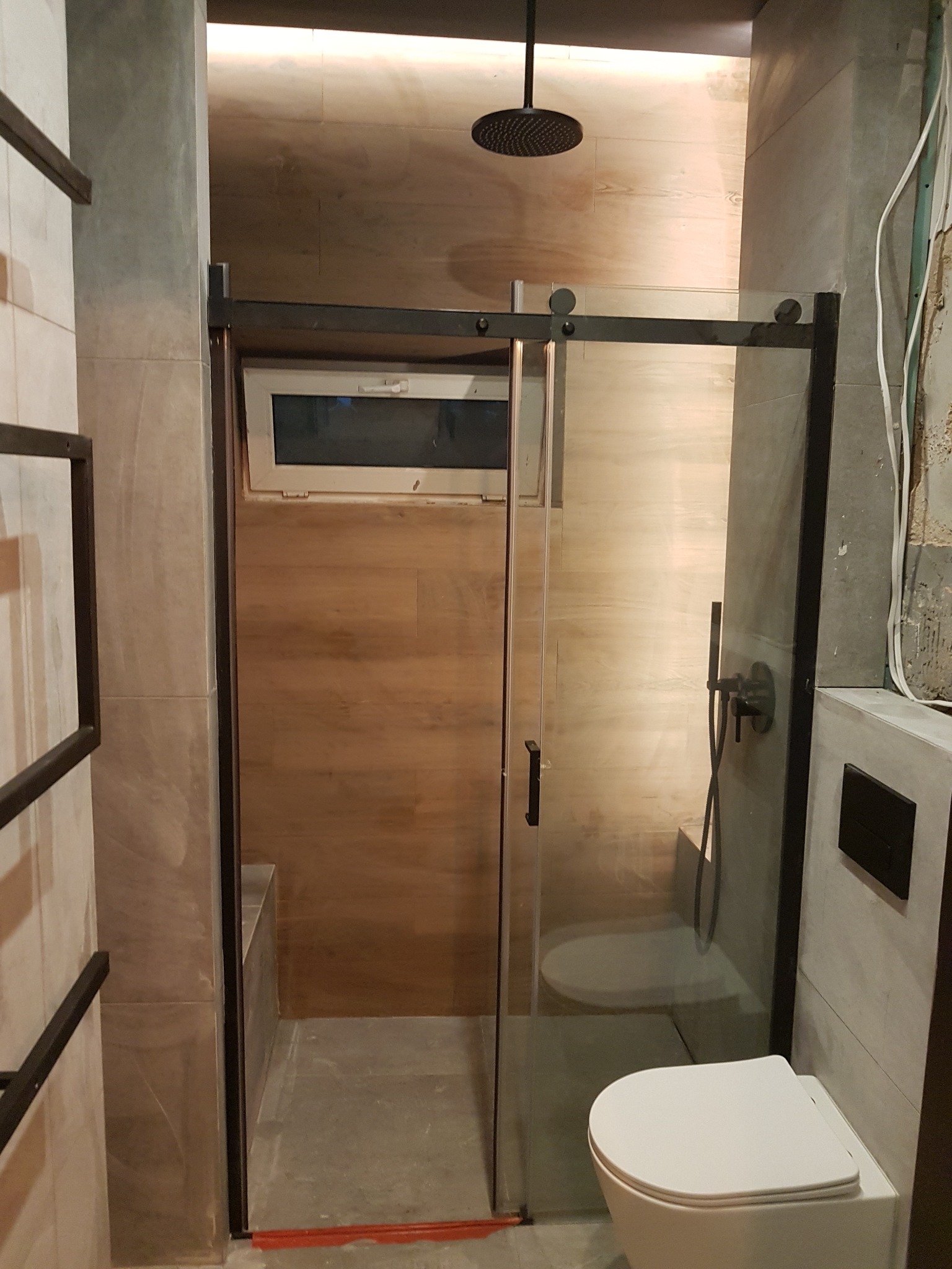 Nowoczesna kabina prysznicowa z przesuwnymi drzwiami w czarnej ramie, widok na zamontowaną toaletę i fragment ściany z niedokończonymi pracami wykończeniowymi.