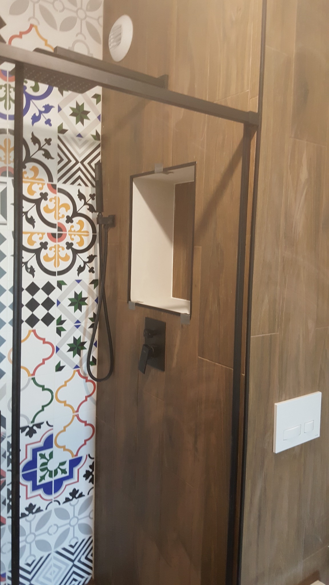 Kabina prysznicowa z czarną ramą, płytki patchworkowe na ścianie, drewnopodobne płytki na pozostałych ścianach, wnęka z półką, czarna bateria prysznicowa i biały panel sterowania toaletą.