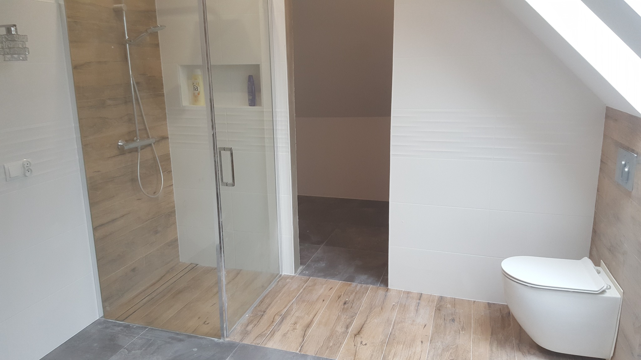 Nowoczesna łazienka na poddaszu z kabiną prysznicową ze szklanymi drzwiami, wiszącą toaletą i drewnopodobnymi płytkami w strefie prysznica i na podłodze, kontrastującymi z szarymi płytkami przy...